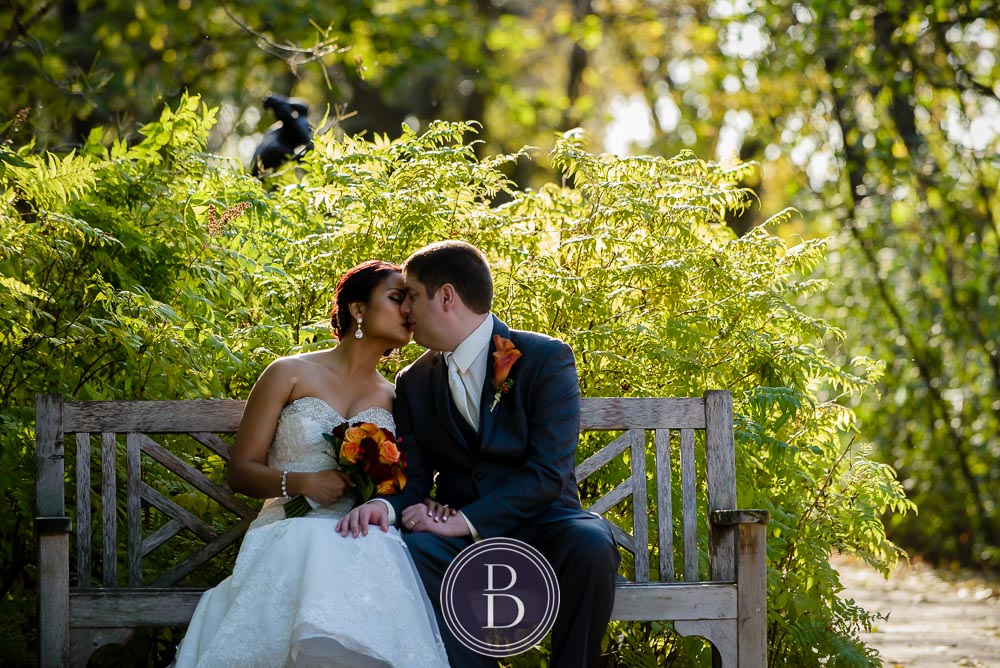 Winnipeg wedding photos portrait groom and bride kissing on a bench at Assiniboine Park surrounded by trees